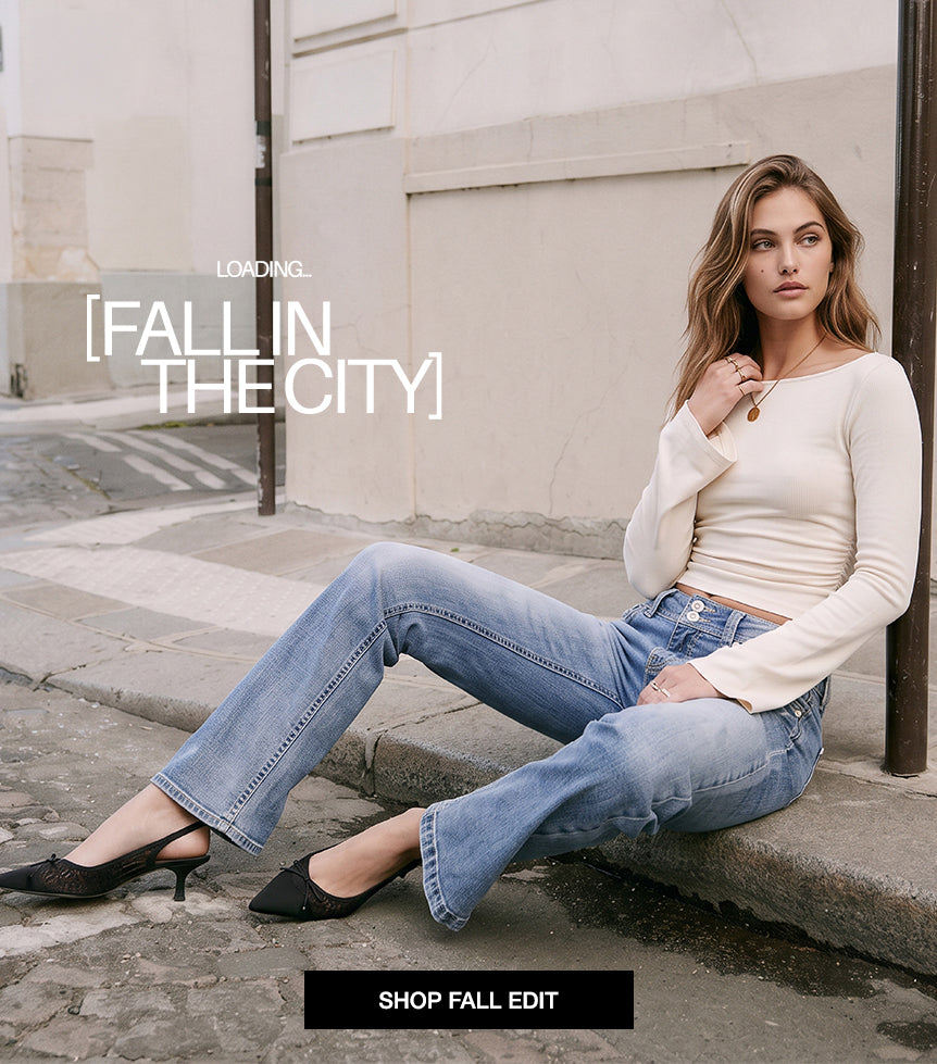 A woman sits on a city sidewalk wearing a fitted cream long-sleeve top, light blue jeans, and black pointed flats. The image includes overlaid text reading 'LOADING: FALL IN THE CITY'.