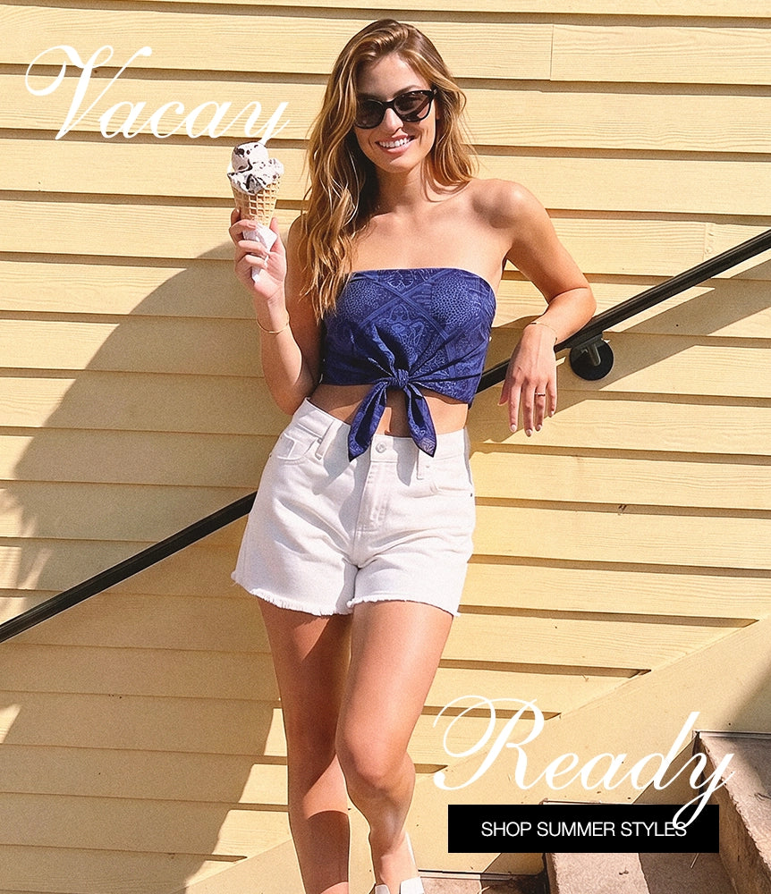 A woman stands outdoors against a beige wooden wall holding an ice cream cone, wearing a navy strapless lace crop top tied at the bust with white denim shorts. Large decorative text overlays read 'Vacay' and 'Ready SHOP SUMMER STYLES'.