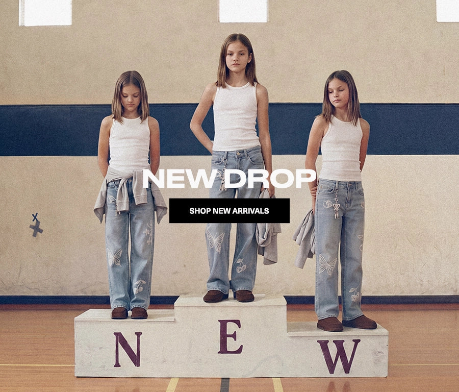 Three girls stand on a podium in a gym wearing white tank tops and distressed light-wash jeans, with large overlaid text reading 'NEW DROP' and 'SHOP NEW ARRIVALS'.