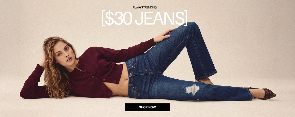 A woman reclines on a beige studio floor wearing blue distressed jeans and a burgundy cropped long-sleeve top, with promotional text reading '$30 JEANS' and a black 'SHOP NOW' button.