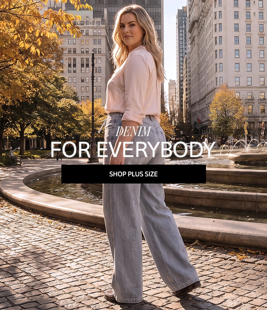 A woman stands in a city plaza wearing light-wash wide-leg denim jeans and a pale pink top, with large overlaid text reading 'DENIM FOR EVERYBODY' and 'SHOP PLUS SIZE'. Trees with yellow autumn leaves and fountain water are visible in the background.