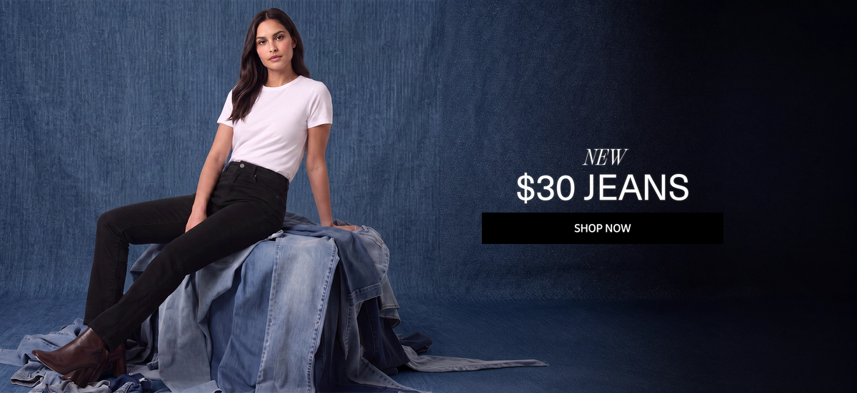 A woman sits on a pile of denim jeans against a dark blue textured backdrop, wearing a white t-shirt and black skinny jeans. Text on the right reads 'NEW $30 JEANS' with a 'SHOP NOW' button.